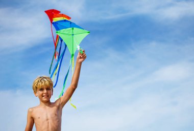 Close-up of a boy holding many kites in hand smiling over blue sky