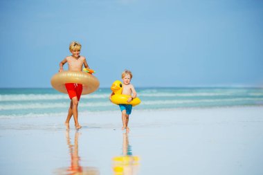 Two happy kids boys with inflatable toy buoys doughnut and duck having fun running on the sand beach