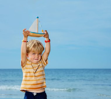 Cute handsome little blond boy hold toy boat playing at the sea beach during summer vacation