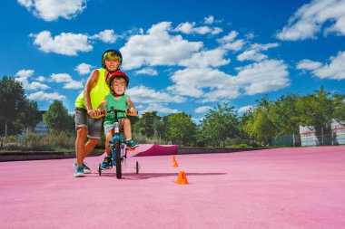 Preteen boy help little brother to learn ride a bicycle pushing the bike with hands in the park going around orange cones
