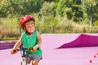 Close-up portrait of a little blond smiling boy ride small bicycle on color surface of the park