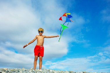 Young smiling boy portrait stand on pebble beach holding many colorful kites in hand over blue sky wearing sunglasses