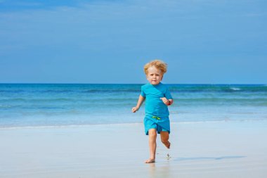 Happy blond little boy run fast on the sand beach over ocean on background