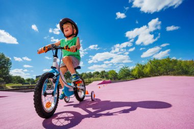 Happy little boy learning to ride bicycle with extra wheels, holds steering wheel and pedals, low angle photo in the skate park