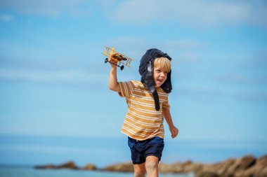 Happy smiling boy run with toy model at the beach of the plane wearing aviation hat and googles