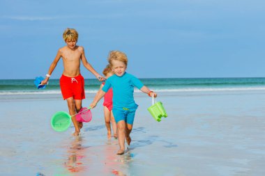 Three kids boys with a girl run from ocean holding buckets and hoop-net having fun, play on a beach at summer