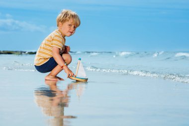 Cute handsome little blond boy put toy boat in the sea waves at the beach during summer vacation