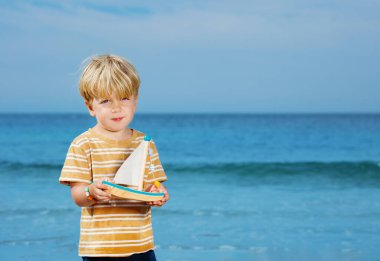 Cute handsome little blond boy pose holding toy boat playing at the sea beach during summer vacation