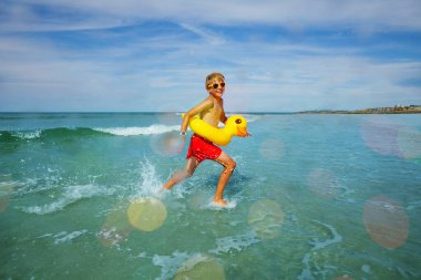Young boy in orange sunglasses run on the beach at the sea with inflatable duck view from side