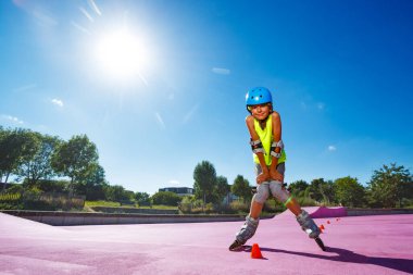 Young boy pose standing in blue helmet skate fast on rollerblades in the skatepark wearing protection and going around cones