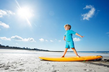 Cute blond boy stand on the surfboard learning balance practicing surfing before going to ocean - view in profile over sun