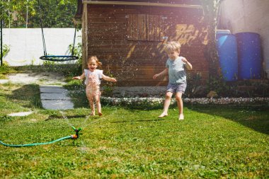 Two happy little children boy and girl run around water sprinkler in the garden lawn on hot summer day