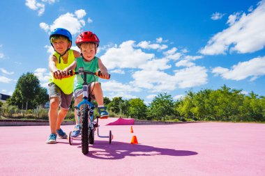 Preteen boy help little brother to learn ride a bicycle pushing the bike with hands in the park