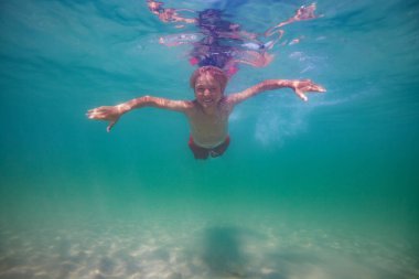 Cute smiling boy swim underwater in the ocean with open eyes and posing over sunlight lit background