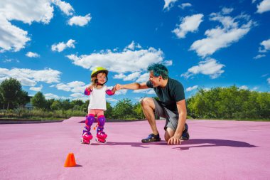 Dad teach little happy girl to stand on rollers holding her with hands at the color skate park