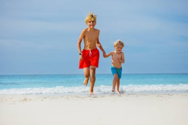 Two children, brothers holding hands running barefoot in sea waves on ocean shore