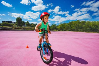 Little blond child ride small bicycle with learning wheels roll around orange cones on color surface of the park
