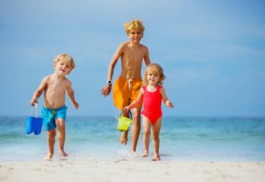 Group kids blond boys and a girl, siblings run barefoot in ocean sand beach carry toy buckets with water