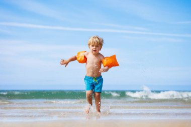 Happy little blond boy run on the ocean beach with waves wearing inflatable shoulder straps