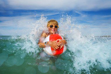 Boy splashing in ocean waves in orange sunglasses with color inflatable ball playing in water