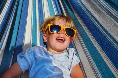 Close-up view from above of a laughing cute little blond boy in sunglasses rest in hammock