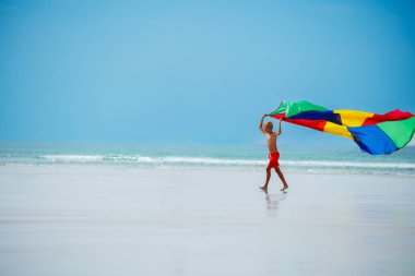 Happy boy run holding colorful parachute fabric flying on the wind on ocean beach side profile view