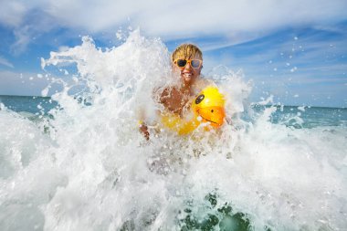 Boy splashing in ocean waves in orange sunglasses with yellow inflatable duck playing in water
