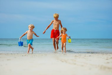 Two boys and a girl, siblings running barefoot in sea waves on ocean shore carry toy buckets with water