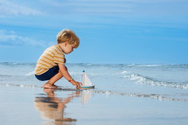 Happy handsome little blond boy put toy boat in the ocean waves at the beach during summer vacation