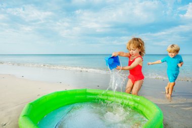 Two happy children run to inflatable pool bringing water from sea in buckets and splash inside