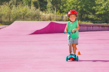 Portrait of the little cute small boy on the kick scooter ride on the skate park with color surface in helmet