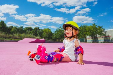 Cute little two years old girl learn to skate on rollers, sit with helmet and protection on the color floor of the park