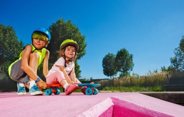 Side view of big brother with little girl sit on the skate at skatepark playing and riding on the ramp