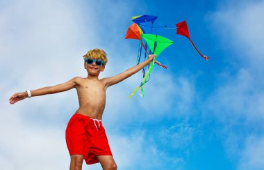 Young smiling boy close-up portrait stand holding many colorful kites in hand over blue sky wearing sunglasses