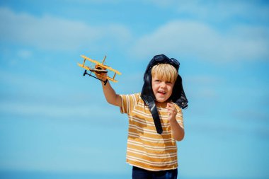 Happy blond boy run with toy model at the beach of the plane wearing aviation hat and googles