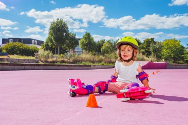 Little girl learn to skate on rollers, sit smiling with helmet and protection on the color floor of the park