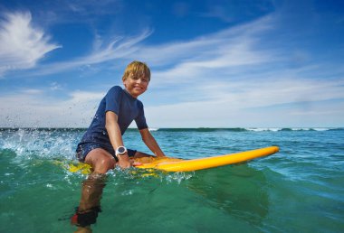 Portrait of smiling boy sit on the surf board looking at camera in the sea learning at surfing school