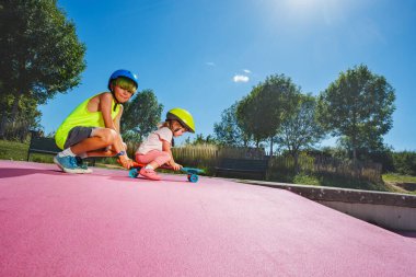 Side view of big brother play with little sister who sits on the skate and wear helmet at skatepark pushing her on the ramp