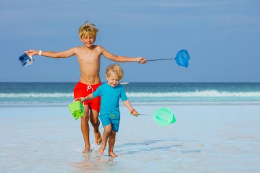 Two young boys brothers run on ocean beach holding buckets and hoop-net having fun