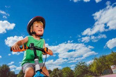 Close-up low angle portrait of a little cute smiling boy ride small bicycle on color surface of the park over blue sky