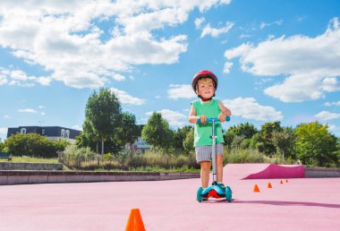 Small blond boy on the kick scooter ride around orange cones on the skate park with color surface in red helmet