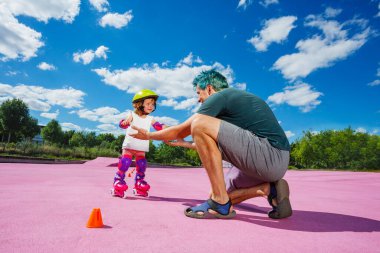 Father teach little happy girl to stand on rollers catching her with hands at the color skate park