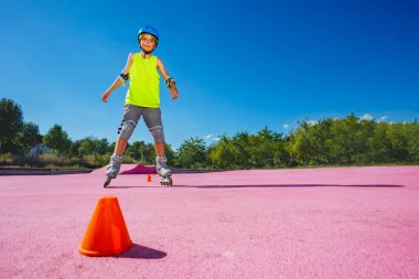 Preteen boy stand in blue helmet with rollerblades in the skatepark wearing protection going fast around orange cones