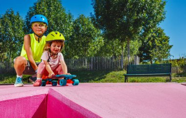 Big brother play with little sister who sits on the skate and wear helmet at skatepark pushing her on the ramp