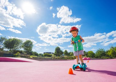 Portrait of the little cute small boy on the kick scooter ride around orange cones on the skatepark with color surface in helmet