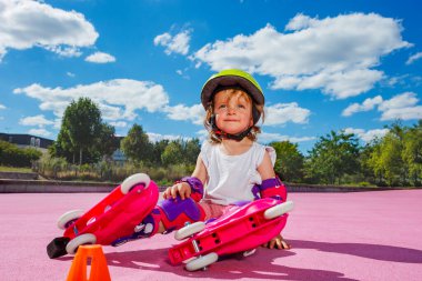 Smiling little one year old girl learn to skate on rollers, sit with helmet and protection on the color floor of the park