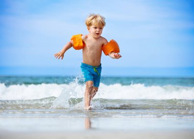 Little blond child boy run on the ocean beach with waves wearing inflatable shoulder straps