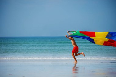 Cute boy run holding colorful parachute fabric flying on the wind on sea beach