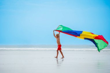 Cute boy walk holding colorful parachute fabric flying on the wind on sea beach