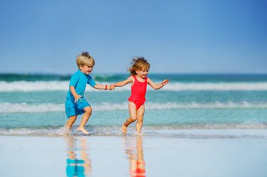 Little boy and girl run on sand beach over ocean waves holding hands smiling having fun profile view